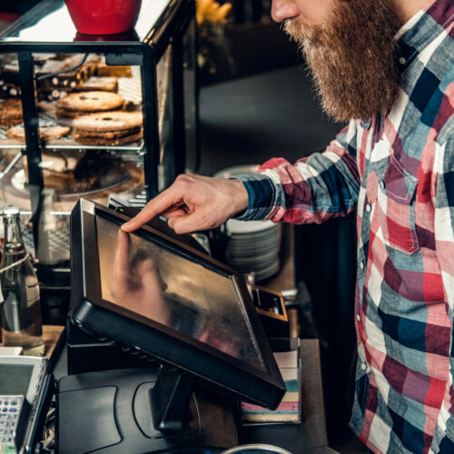 A man at the counter using cash register in a coffee shop. Positive bearded male at the counter using cash register in a coffee shop.